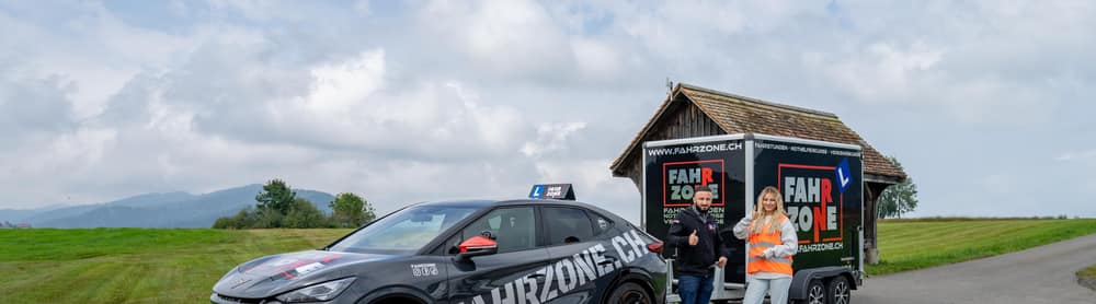 Zwei Personen stehen neben einem schwarzen Auto und einem Anhänger mit der Aufschrift FAHRZONE.CH.