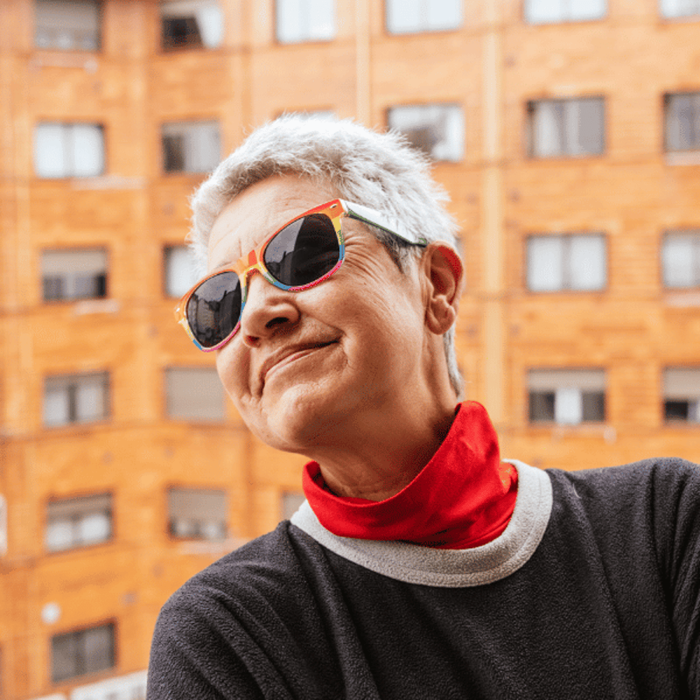 Eine lächelnde Person mit kurzem grauen Haar und bunter Sonnenbrille vor einem orangefarbenen Gebäude.
