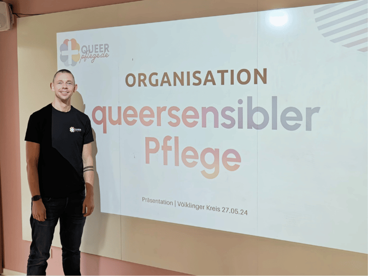 Lächelnder Mann vor einer Präsentation „Organisation queersensibler Pflege“.