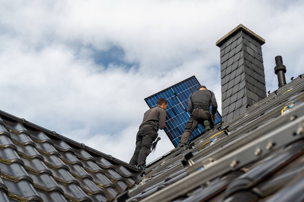 Zwei OurSun-Monteure tragen ein Solarmodul über ein graues Ziegeldach