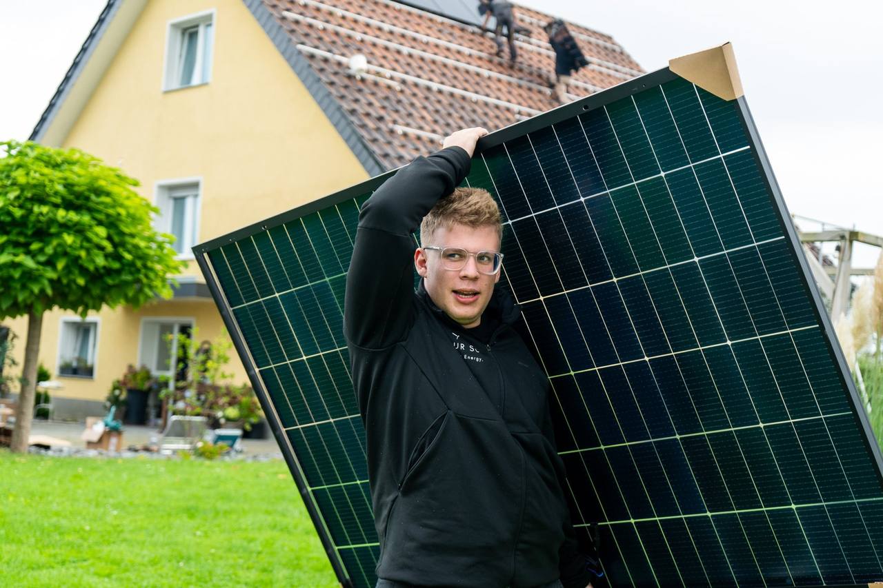 OurSun-Mitarbeiter trägt ein Solarmodul, im Hintergrund die Montage auf dem Dach