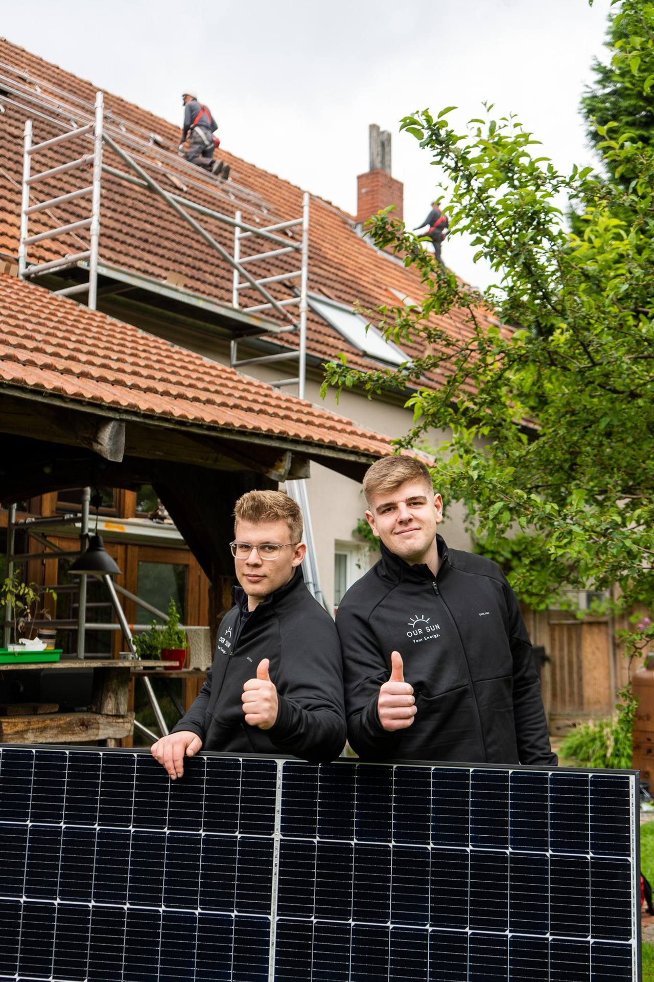 Zwei OurSun-Mitarbeiter zeigen den Daumen hoch hinter einem Solarmodul