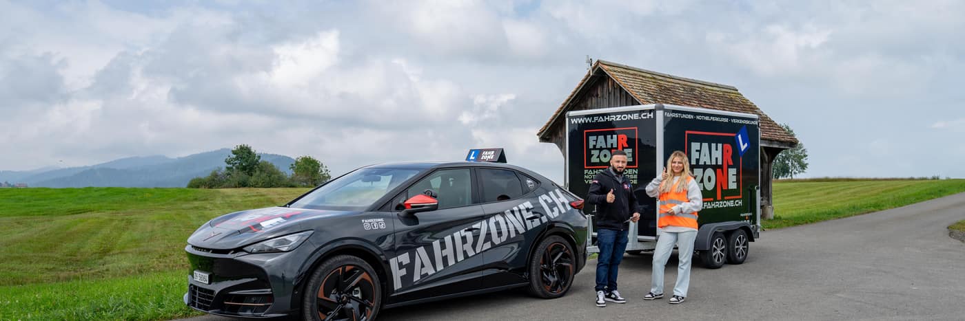 Schwarzes Elektroauto mit FAHRZONE.CH-Schriftzug und Fahrschulschild, dahinter ein Anhänger.