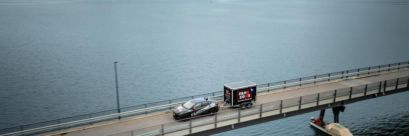 Ein FAHRZONE-Auto mit Anhänger fährt über eine Brücke über einen See.