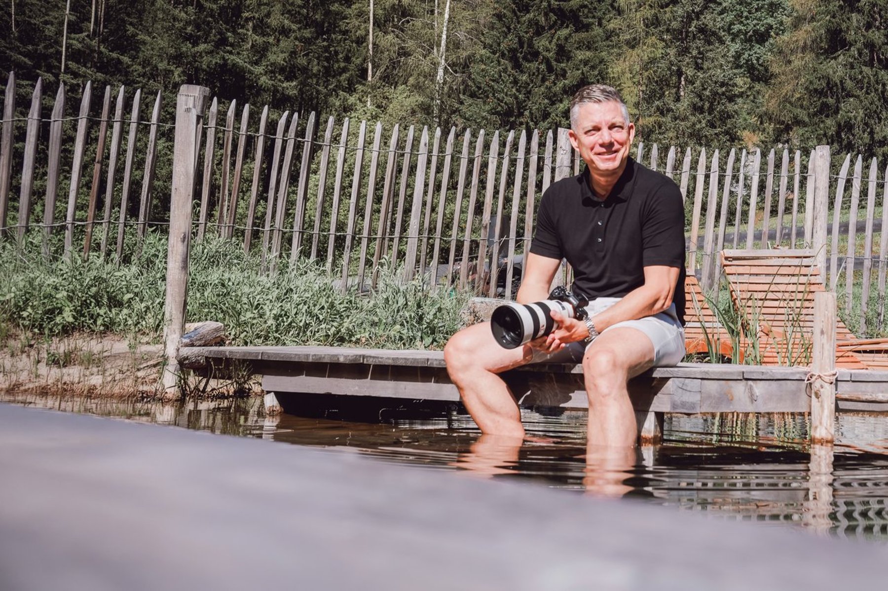 Alex Lörtscher sitzt lachend mit seiner Kamera auf einem Holzsteg am Wasser