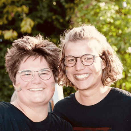 Zwei lächelnde Personen mit aufgemalten Regenbogenfarben im Gesicht.