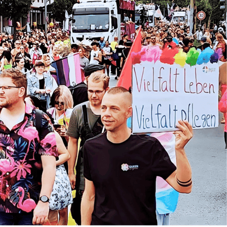 Person mit Schild „Vielfalt leben, Vielfalt pflegen“ auf einer Pride-Parade.