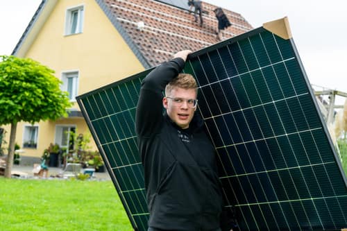 OurSun-Mitarbeiter trägt ein Solarmodul, im Hintergrund die Montage auf dem Dach
