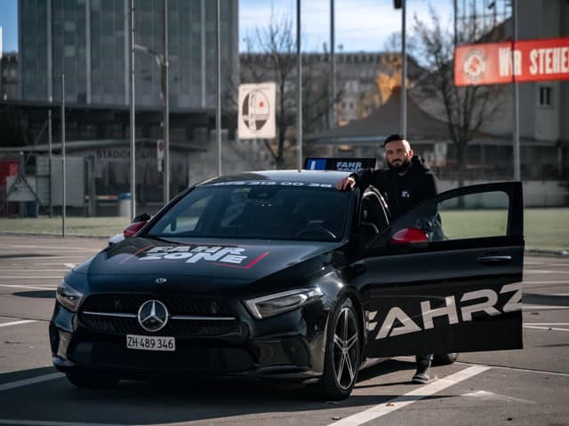 Fahrlehrer der FAHRZONE GmbH steht am schwarzen Fahrschulauto.