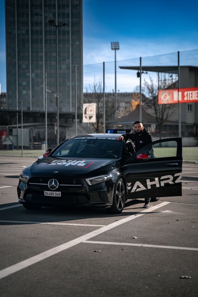 Ein Fahrlehrer lehnt an einem schwarzen FAHRZONE-Auto in der Stadt.