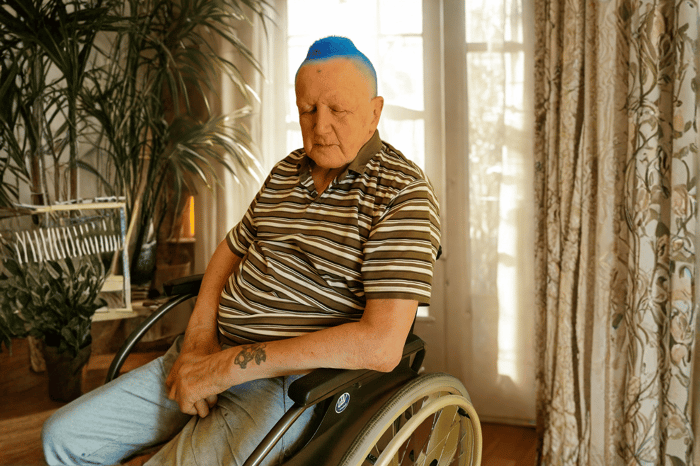 Älterer Mann mit blauen Haaren und gestreiftem Hemd in einem sonnigen Raum.