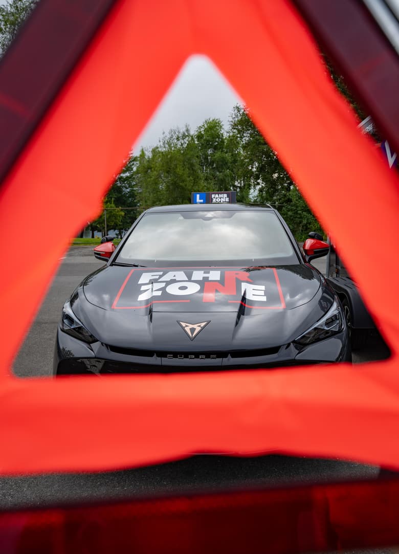 Schwarzes Auto mit FAHRZONE-Schriftzug auf der Motorhaube, gesehen durch ein rotes Warndreieck.