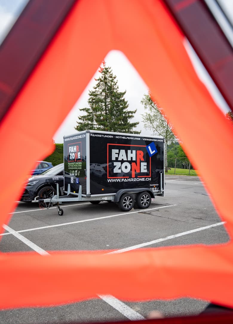 Schwarzer Anhänger mit FAHRZONE-Schriftzug und L-Schild, gesehen durch ein rotes Warndreieck.