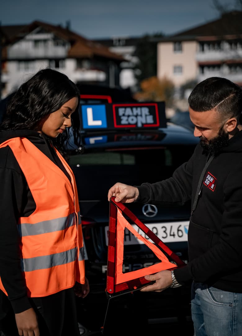 Fahrlehrer zeigt einer Fahrschülerin ein Warndreieck hinter einem schwarzen Auto mit FAHRZONE-Schild.