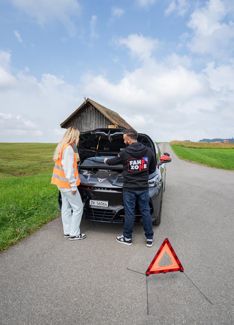 Fahrlehrer und Fahrschülerin schauen gemeinsam in den Motorraum eines schwarzen Autos, davor ein Warndreieck.