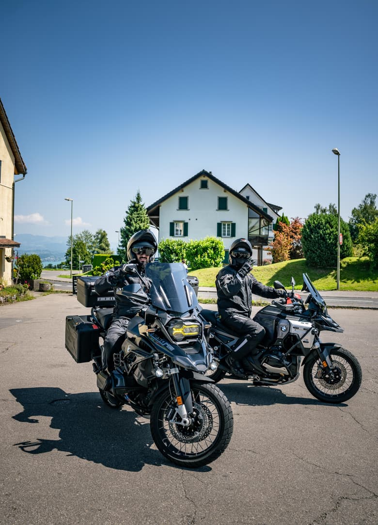 Zwei Motorradfahrer in voller Montur auf dunklen Motorrädern stehen auf einer Strasse.