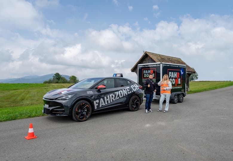 Schwarzes Elektroauto mit FAHRZONE.CH-Schriftzug und Fahrschulschild, dahinter ein Anhänger.