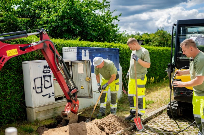 Ein Mitarbeiter schachtet von Hand neben einem Verteilerschrank, dahinter ein Bagger.