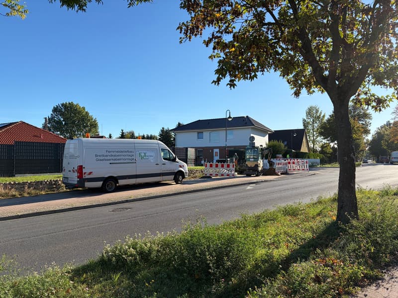 Weißer Transporter mit FKT-Aufschrift am Straßenrand, dahinter ein Minibagger und rot-weiße Absperrschranken vor einem Wohnhaus.