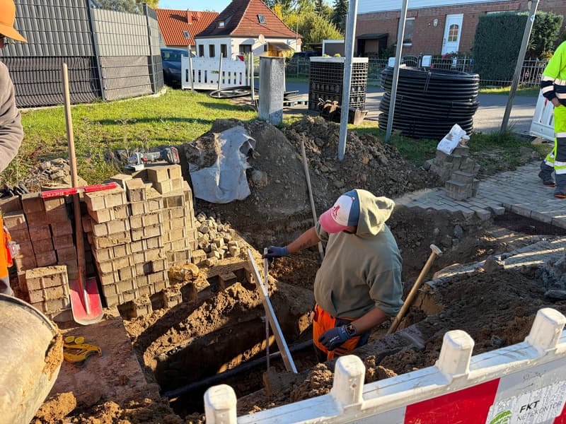 Mitarbeiter im Graben hält einen Zollstock neben eine Richtlatte, auf der Grabensohle liegt ein schwarzes Rohr.