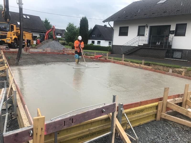Mitarbeiter glättet den frischen Beton einer Bodenplatte, dahinter die Betonpumpe
