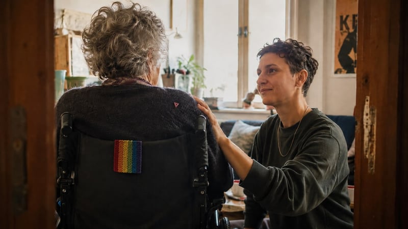 Pflegende Person legt die Hand auf die Schulter einer älteren Person im Rollstuhl mit Regenbogen-Aufnäher.