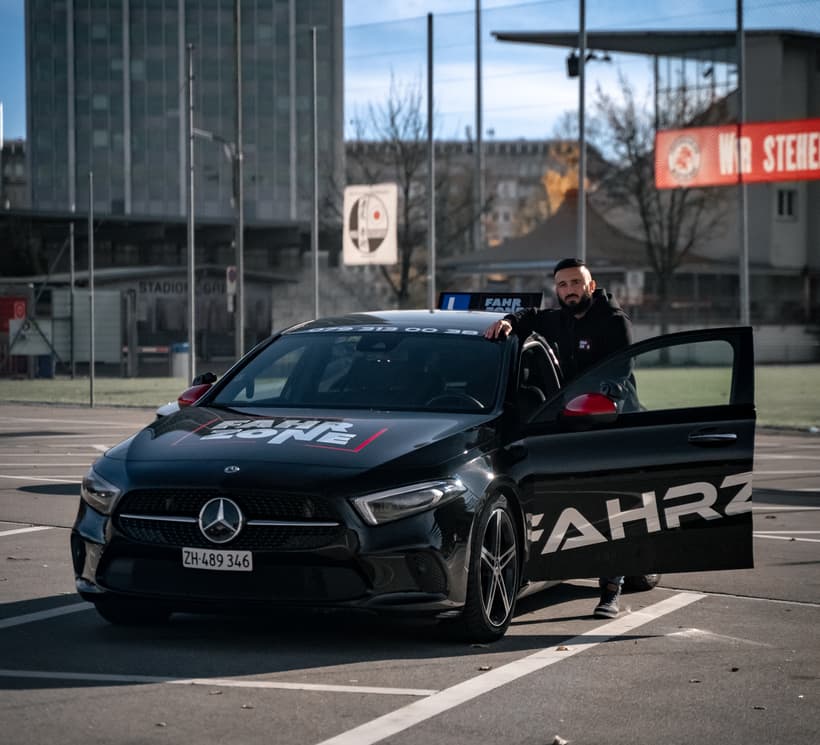 Schwarzes FAHRZONE-Fahrzeug mit geöffneter Fahrertür und einer Person daneben auf einem Parkplatz.
