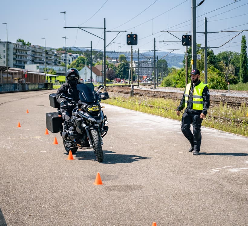 Motorradfahrer fährt durch einen Parcours aus orangefarbenen Kegeln, daneben ein Instruktor.
