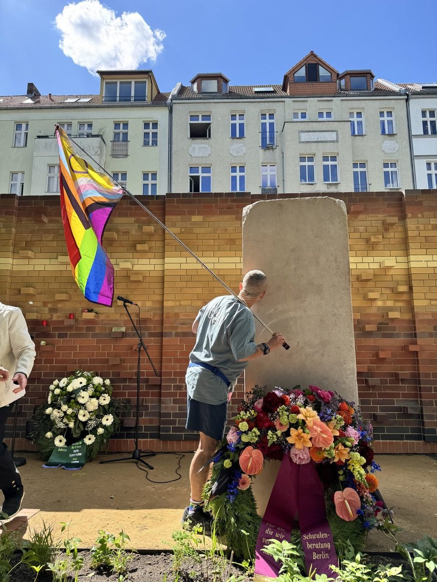 Person spricht am Rednerpult bei einer queeren Kundgebung im Freien, mit Progress-Pride-Flagge und Blumengestecken.