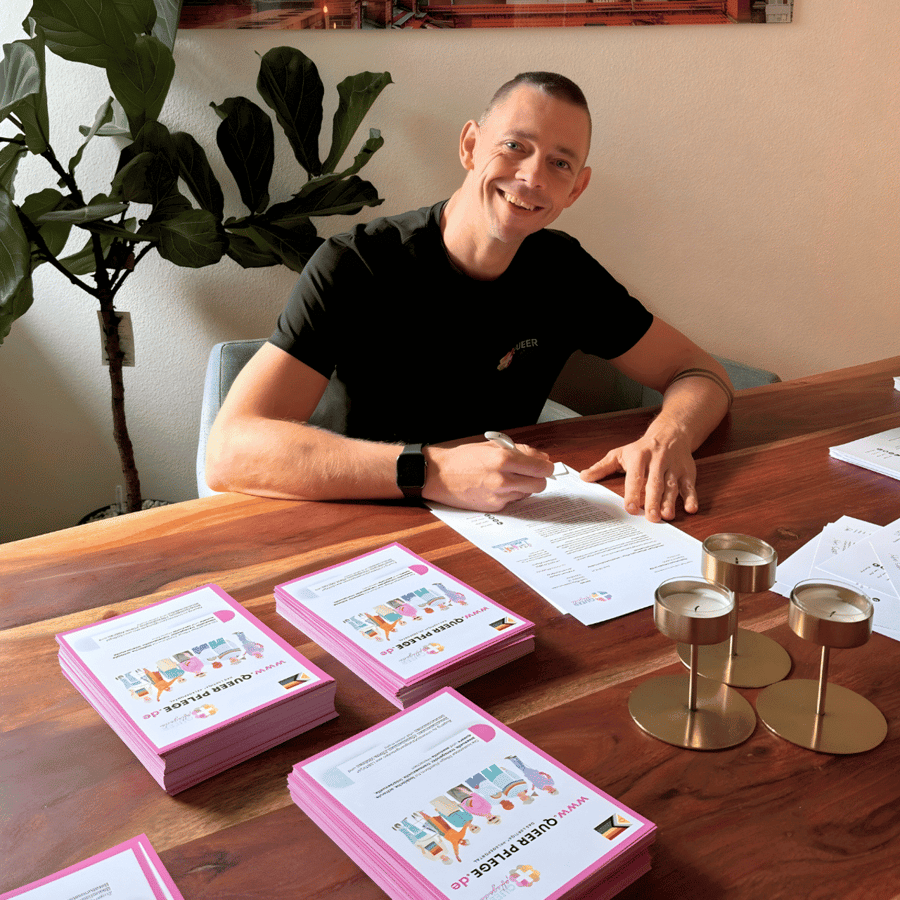 Andreas an einem Tisch mit Stapeln pinker QueerPflege-Flyer.