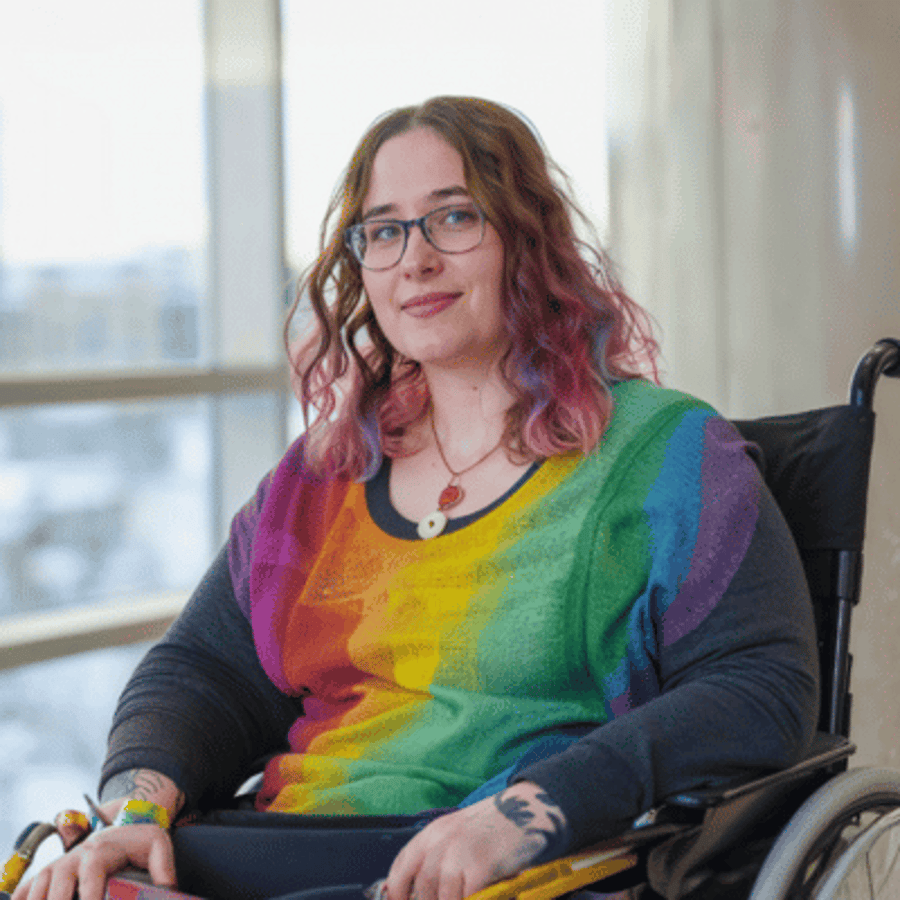 Junge Person im Rollstuhl mit Brille, bunten Haaren und Regenbogen-Pullover lächelt.