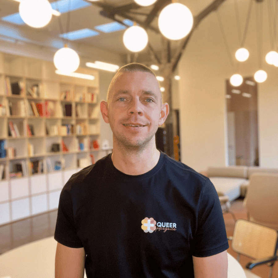 Lächelnder Mann mit QueerPflege-Shirt in einem Raum mit Bücherregalen.