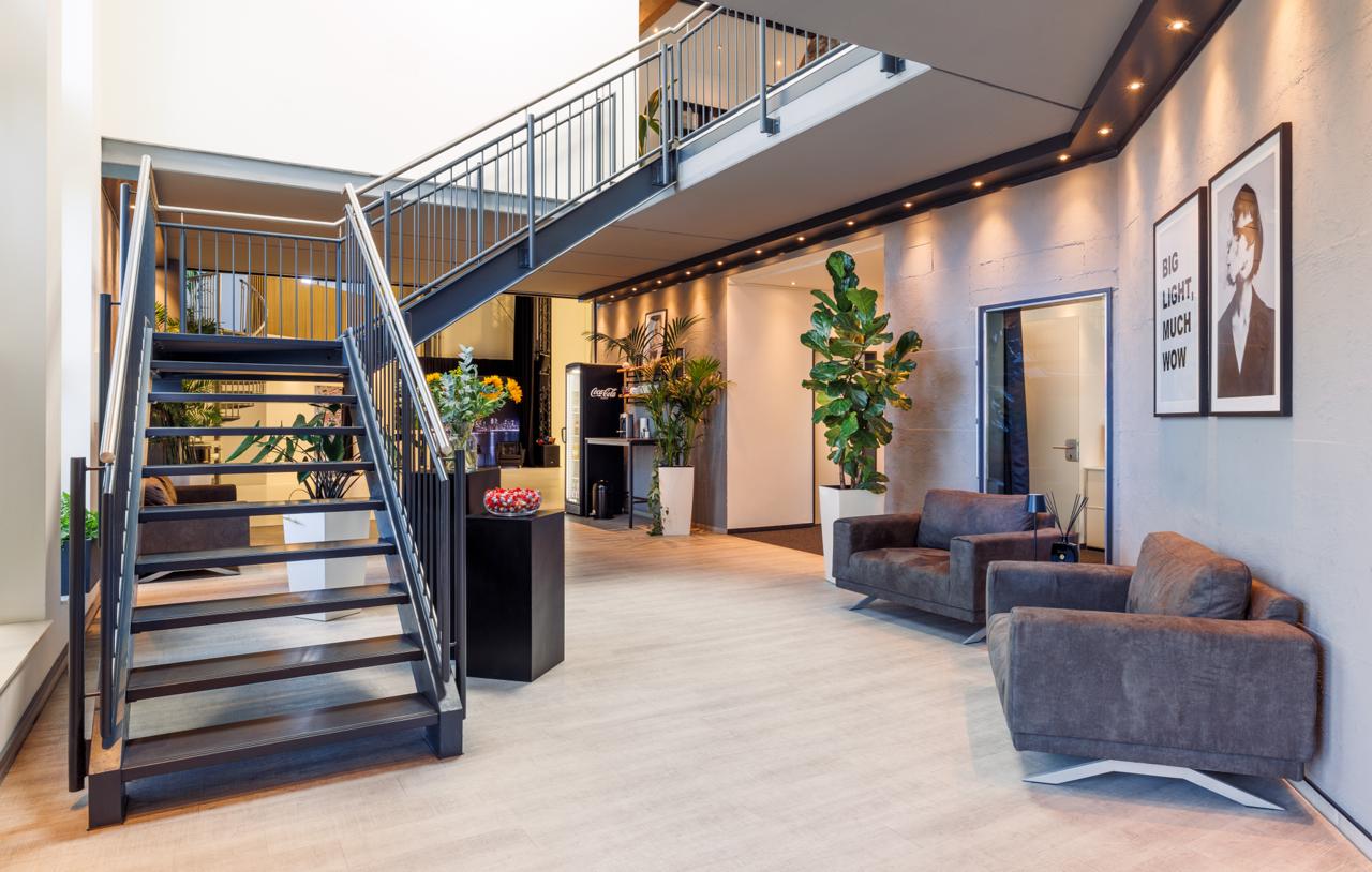 Bürolounge mit offener Stahltreppe, zwei grauen Sesseln und Pflanzen