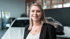 Eine lächelnde blonde Frau im schwarzen Blazer steht in einem Autohaus.