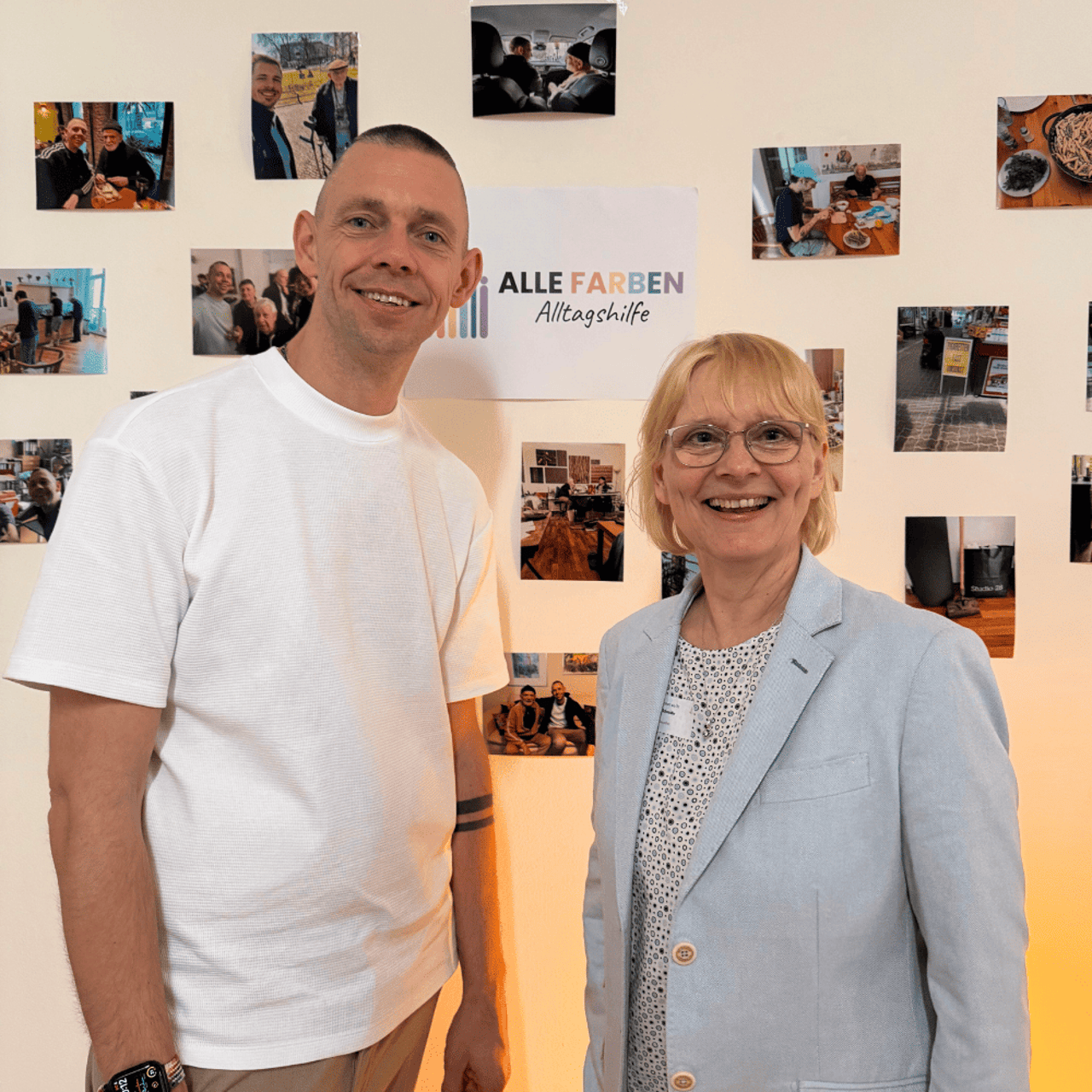 Ein Mann und eine Frau lächeln vor einer Fotowand; ein Logo "ALLE FARBEN Alltagshilfe" ist sichtbar.