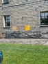 Graue Natursteinmauer vor einer Schieferfassade mit Pflasterweg und Rasenfläche im Vordergrund.