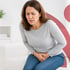Woman in grey shirt and jeans sitting on a white couch, clutching her stomach in pain.