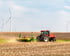 Roter Traktor pflügt ein Feld unter blauem Himmel mit weißen Windturbinen im Hintergrund.