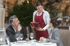 A smiling waitress in a white shirt and red apron takes an order from a man and a woman at a restaurant table.