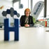 Frau sitzt an einem Konferenztisch mit einem blauen "H" im Vordergrund und einem "Herrscher" Banner im Hintergrund.