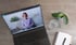 Laptop mit Videokonferenz einer lächelnden Frau, daneben eine Pflanze, Wasserglas und Notizbuch.