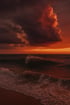 Dunkle Wellen schlagen gegen den Sandstrand unter einem feurigen, rot-orangefarbenen Sonnenuntergangshimmel.