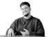 Ein lächelnder junger Mann mit Bart und dunklem Pullover sitzt an einem Tisch und gestikuliert mit der Hand.