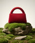 Rote flauschige Handtasche auf einem bemoosten Stein, umgeben von Moos und kleinen Steinen.