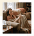A woman sits on a sofa wrapped in a blanket next to a box of tissues and a glass of water.