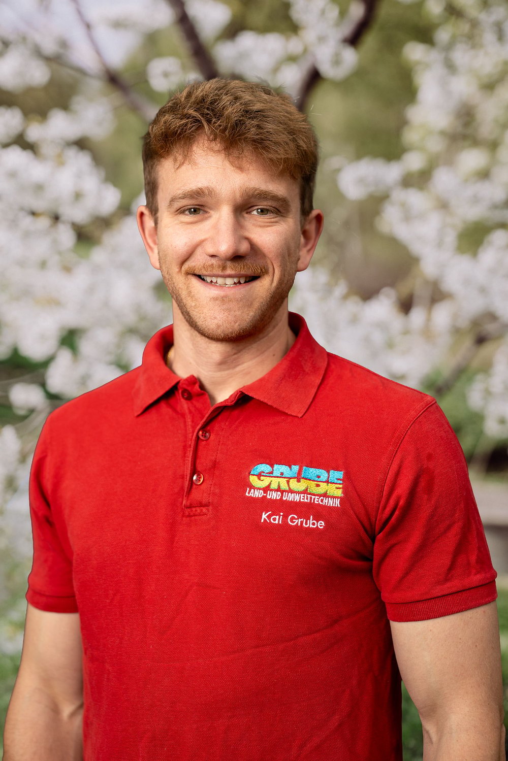 Frau mit grauen Haaren und Brille in blauem Poloshirt mit "GRÜN" Logo vor weiß blühendem Baum.