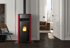 Roter Pelletkamin mit schwarzem Rauchrohr und sichtbarer Flamme in einem Wohnzimmer.