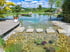 Ein großer Schwimmteich mit Trittsteinen im klaren Wasser, Holzterrasse und grüner Gartenlandschaft unter blauem Himmel.