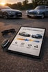 Tablet mit "AN- & VERKAUFSBERATUNG" und zwei Autos, Sonnenbrille, Autoschlüssel auf Asphalt vor zwei Autos.