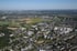 Luftaufnahme einer Stadt mit zahlreichen Gebäuden, Grünflächen, Feldern und einem Sportstadion unter blauem Himmel.